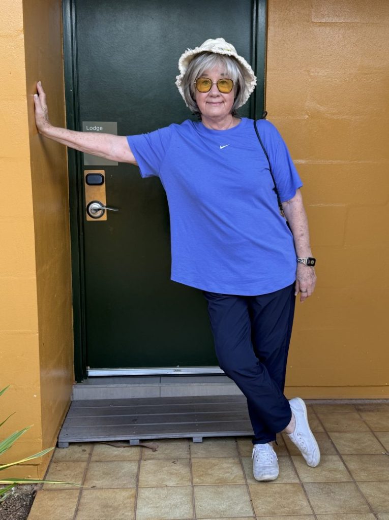 An older woman stands by a green door wearing a blue t-shirt and navy pants, with a white hat and yellow sunglasses.